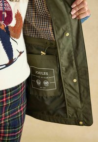 Zielona wodoodporna kurtka z w kratkę wewnętrzną. Posiada kieszeń z logo oraz sznurek do regulacji. Złote akcenty na dodatkowych elementach.