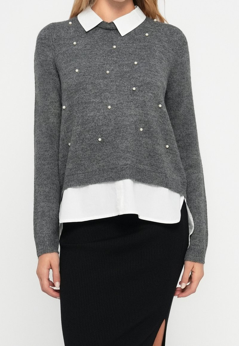 Femme portant un pull gris avec des perles éparses sur une chemise blanche à col, associée à une jupe noire côtelée avec une fente latérale.