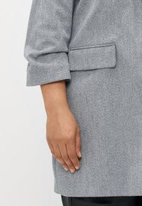 Grijze herringbone-patroon stofjas met een opgerolde manchette, voorzien van een voorzak. Gladde textuur en getailleerde pasvorm.
