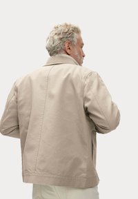 Beige katoenen jas met een geribde kraag, zijzakken en een ontspannen pasvorm. De achterkant heeft een centrale naad. De textuur lijkt glad te zijn.