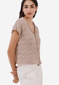Knappt kortärmad cardigan i en ljusbeige färg, med ett texturerat spetsmönster och knäppning framtill. Sidoklipp är synliga.