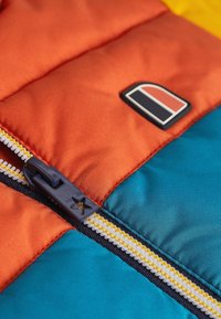 Polstret jakke med sektioner i orange, teal og gul. Har en lynlåslukning med en marineblå trækflap og et logo patch. Teksturert stof.