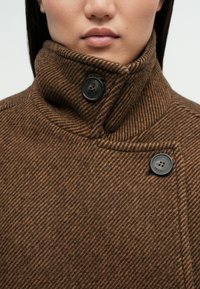 Καφέ, ριγέ ύφασμα σακάκι με υψηλό γιακά, δύο μαύρα κουμπιά και λείες υφές, που τονίζουν τα σύγχρονα σχεδιαστικά στοιχεία.