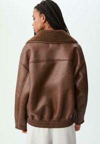 Persona con capelli intrecciati che indossa grandi orecchini a cerchio in oro e un giubbotto di pelle marrone oversize con colletto in pelliccia, voltata di spalle.