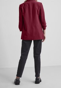 Maroon blazer med en strukturerad design, långa ärmar som är rullade vid manschetterna, parat med mörkgrå slim-fit byxor och kraftiga svarta skor.