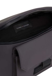 Bolso sintético gris con cierre de solapa magnético, bolsillo interior con cremallera y una etiqueta que dice "TOMMY HILFIGER". Textura suave.