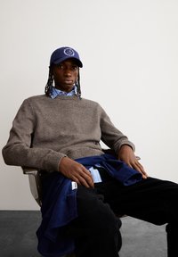 Brauner Strickpullover über einem blau gestreiften Hemd, dunkle Cordhosen und eine marineblaue Mütze mit einem Logo, sitzt auf einem Stuhl.