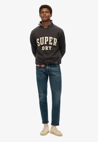 Sudadera gris oscuro con "SUPER DRY" en letras crema, superpuesta a una camisa de cuadros, combinada con jeans azules y zapatillas beige.