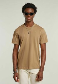 T-shirt marrone a maniche corte in una vestibilità rilassata, realizzato in tessuto morbido con colletto rotondo. Presenta un piccolo logo sul petto e un'etichetta laterale.