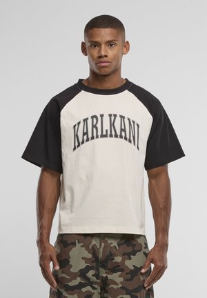 Tânăr purtând un tricou alb și negru cu mâneci raglan, cu textul „KARLKANI” și pantaloni de camuflaj, stând în fața unui fundal gri simplu.