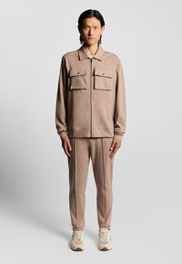 Beige zip-up jacka med två bröstfickor, kombinerad med matchande byxor. Tyget verkar mjukt, med en slät textur och avslappnad passform.