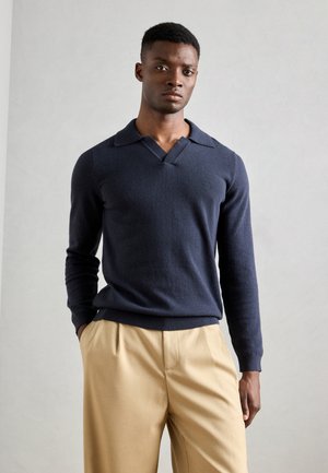 Navy blue sweater with a V-neck and pointed collar, made of soft material. Paired with beige pleated trousers for a neutral contrast.