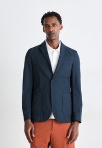 Sacou texturat de culoare albastru marin, cu rever la cută, închidere cu un singur buton și două buzunare frontale, asortat cu un tricou polo alb și pantaloni portocalii.
