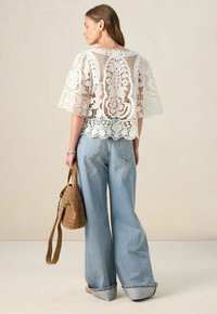 Spitzenoberteil mit weiten Ärmeln, versehen mit floralen Mustern und einem gewellten Saum, kombiniert mit hoch taillierten, hellblauen Jeans und einer beige durchlöcherten Tasche.