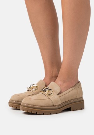 Mocassini in suede beige con suola in gomma spessa, caratterizzati da un vistoso dettaglio in metallo dorato sulla tomaia e cuciture decorative.