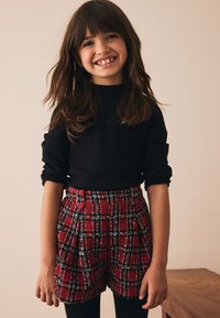 Schwarzes, geripptes Langarmoberteil kombiniert mit rotem karierten Shorts mit strukturierter Musterung. Das Kind hat lange braune Haare und lächelt.