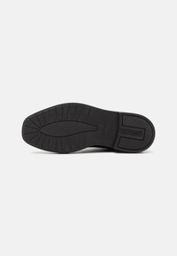 Semelle de chaussure en caoutchouc noir avec un motif de bande de roulement texturé et le nom de la marque embossé près du talon sur un fond blanc uni.