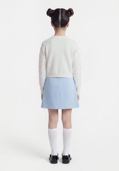 Κορίτσι με χτένισμα διπλό κότσο, φοράει λευκό πουλόβερ με διακοσμημένα μανίκια, ανοιχτό μπλε φούστα, λευκές κάλτσες μέχρι το γόνατο και μαύρα παπούτσια, στραμμένη με την πλάτη.