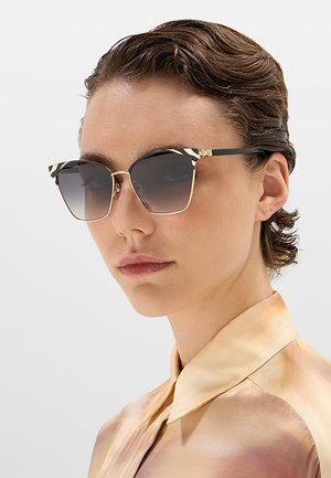 Persona con capelli castani corti indossa occhiali da sole quadrati sfumati con montatura decorata nera e oro e una camicia beige setosa con collo.