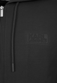 Svart zip-up hoodie med en texturerad lapp som visar "KARL LAGERFELD"-logotypen, med ett slätt material och metalldragkedja.