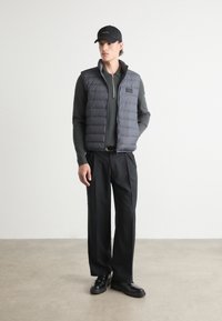 Dunkelgraue Steppweste mit Reißverschluss vorne, kombiniert mit einem dunklen Pullover und schwarzen Hosen. Eine schwarze Mütze rundet das Outfit ab. Glatte Texturen.