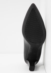 Chaussure à talon haut noire à bout pointu, positionnée avec la semelle vers le haut, présentant une semelle texturée avec un motif répétitif de logo "MK" et un talon bloc.