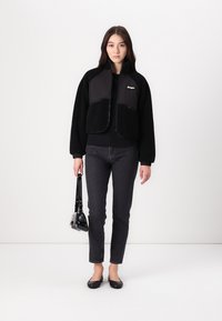 Giacca in pile nera con anteriore imbottito, chiusura con zip e tasche laterali, abbinata a jeans di denim scuro e ballerine nere, mentre tiene una borsa lucida.
