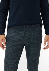 Donkere geruite broeken van gestructureerde stof hebben een subtiel ruitpatroon in tinten van marineblauw en grijs, met twee zijzakken en een knoopsluiting.