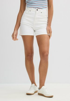 Persona vistiendo pantalones cortos de mezclilla blancos de cintura alta, zapatillas blancas y una blusa sin mangas de rayas, de pie contra un fondo claro.
