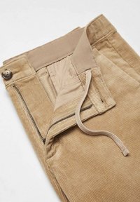 Pantalon beige en velours côtelé avec une fermeture éclair ouverte, fermeture par bouton et cordon visible sur un fond clair.