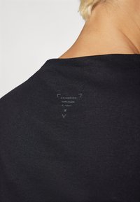 Svart t-skjorte med en glatt tekstur, med en subtil grafikk og logo trykt på baksiden nær halsen, som viser et minimalistisk design.