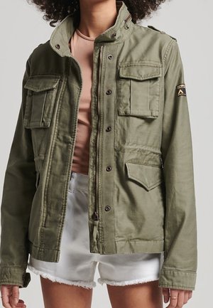 Person iført en åben olivengrøn militærinspireret jakke over en lyserød top med hvide shorts med frynset kant mod en ensfarvet baggrund.