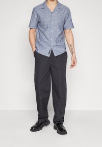 Kurzärmliges, blaues Texthemd mit Knopfleiste und Frontlogo, kombiniert mit schwarzen, locker sitzenden Hosen und schwarzen Schuhen.