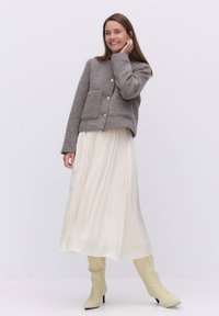 Harmaa villasekoitteinen lyhyt takki, jossa on kaksi etutaskua ja nappisuljenta, yhdistettynä valuvaan ivoriin värjättyyn laskostettuun hameeseen ja polvipitkiin beigen värisiin saappaisiin.