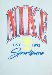 Šviesiai mėlyna marškinėliai su ryškiu raudonu "NIKE" tekstu, geltonu trofėjaus grafiku ir mėlynu "Sportswear" užrašu, turintys retro dizaino stilių.
