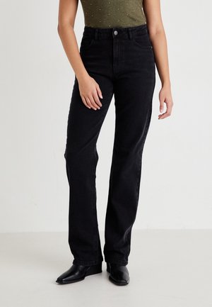 Femme portant un jean noir taille haute à jambes droites et des bottes noires à bout pointu, debout devant un fond clair et uni.