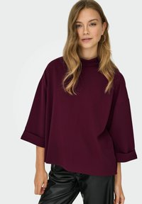 Top oversize bordeaux con collo alto e maniche ampie, realizzato in tessuto morbido. Presenta una vestibilità rilassata e dettagli di cuciture minimi.
