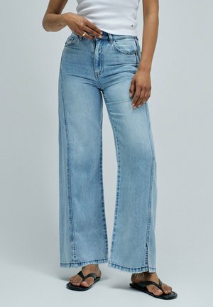 Personne portant un jean bleu clair à jambes larges, un top blanc sans manches et des sandales noires, ajustant la taille d'une main.