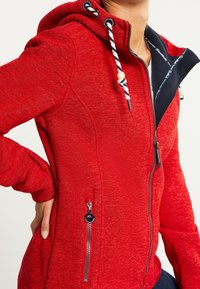 Femme portant un sweat à capuche rouge avec doublure en polaire bleu marine et cordons blanc et bleu marine, la main reposant sur sa hanche.