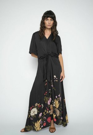 Žena u crnoj maksi haljini s vezanim prednjim dijelom i višebojnim cvjetnim uzorcima blizu ruba, stoji nasuprot jednobojnoj svijetloj pozadini.