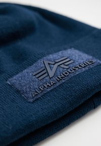Tamna plava pletena kapa s teksturiranim tkanim flasterom s utisnutom oznakom "ALPHA INDUSTRIES" i logotipom s krilima.