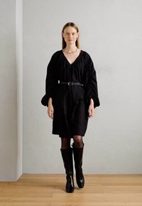 Kobieta idąca do przodu w czarnej sukience z paskiem, o obszernej rękawach, czarnych kozakach do kolan i złotej naszyjniku, stojąca przed gładką ścianą.