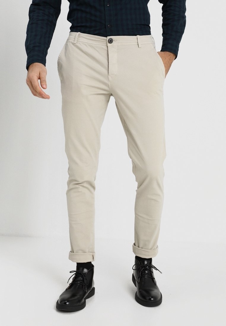 Beige cotton trousers with slim fit, front pockets, and cuffed hems; paired with black lace-up boots.