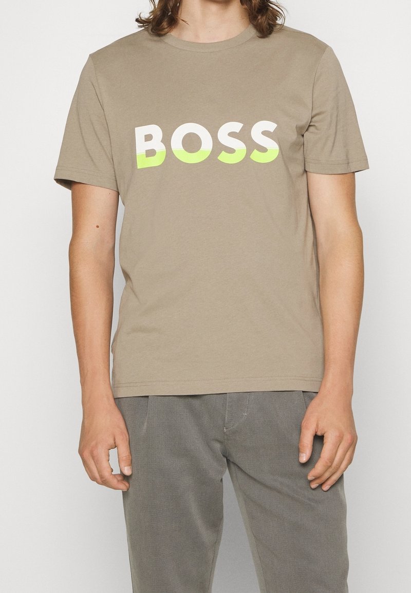 Personne portant un t-shirt beige avec le mot "BOSS" en grandes lettres blanches et vert clair sur la poitrine et un pantalon gris.