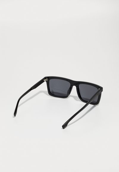 Lunettes de soleil noires rectangulaires avec des verres foncés, monture en plastique brillant et accents latéraux texturés. Des bras fins noirs s'étendent de la monture.