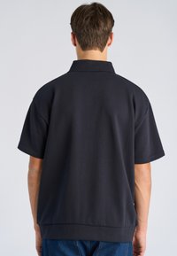 Persoon van achteren gezien, gekleed in een loszittend zwart overhemd met korte mouwen en een kraag, en een blauwe spijkerbroek, tegen een effen achtergrond.