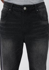 Jeans de mezclilla negra con un diseño clásico de cinco bolsillos, cremallera y cierre de botón metálico. El sutil desgastado agrega textura al material.