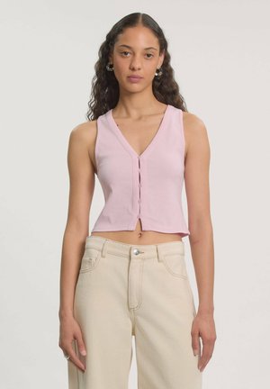 Vrouw met lang krullend haar draagt een roze mouwloze, met knopen gesloten cropped top en een beige hoge taille broek, staand tegen een effen lichte achtergrond.