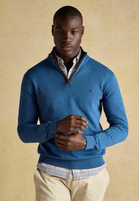 Zils adījuma džemperis ar rāvējslēdzēja apkakli, kombinēts ar rakstu krekliņu ar apkakli zem tā; modelis stāv ar saliktām rokām.