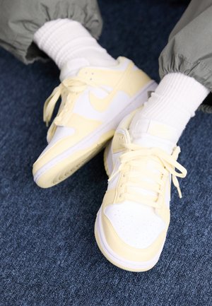 Stopy w białych i jasnożółtych butach sportowych Nike z białymi skarpetkami i zielonymi spodniami na niebieskim, pokrytym dywanem podłożu.